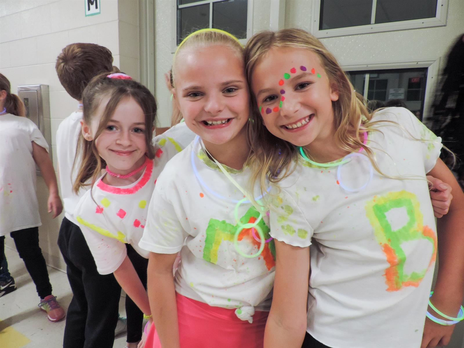 Three girls smiling with white shirts that have glowing paint on it.